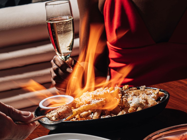 A meal with flames over it with a person in the background holding a wine glass