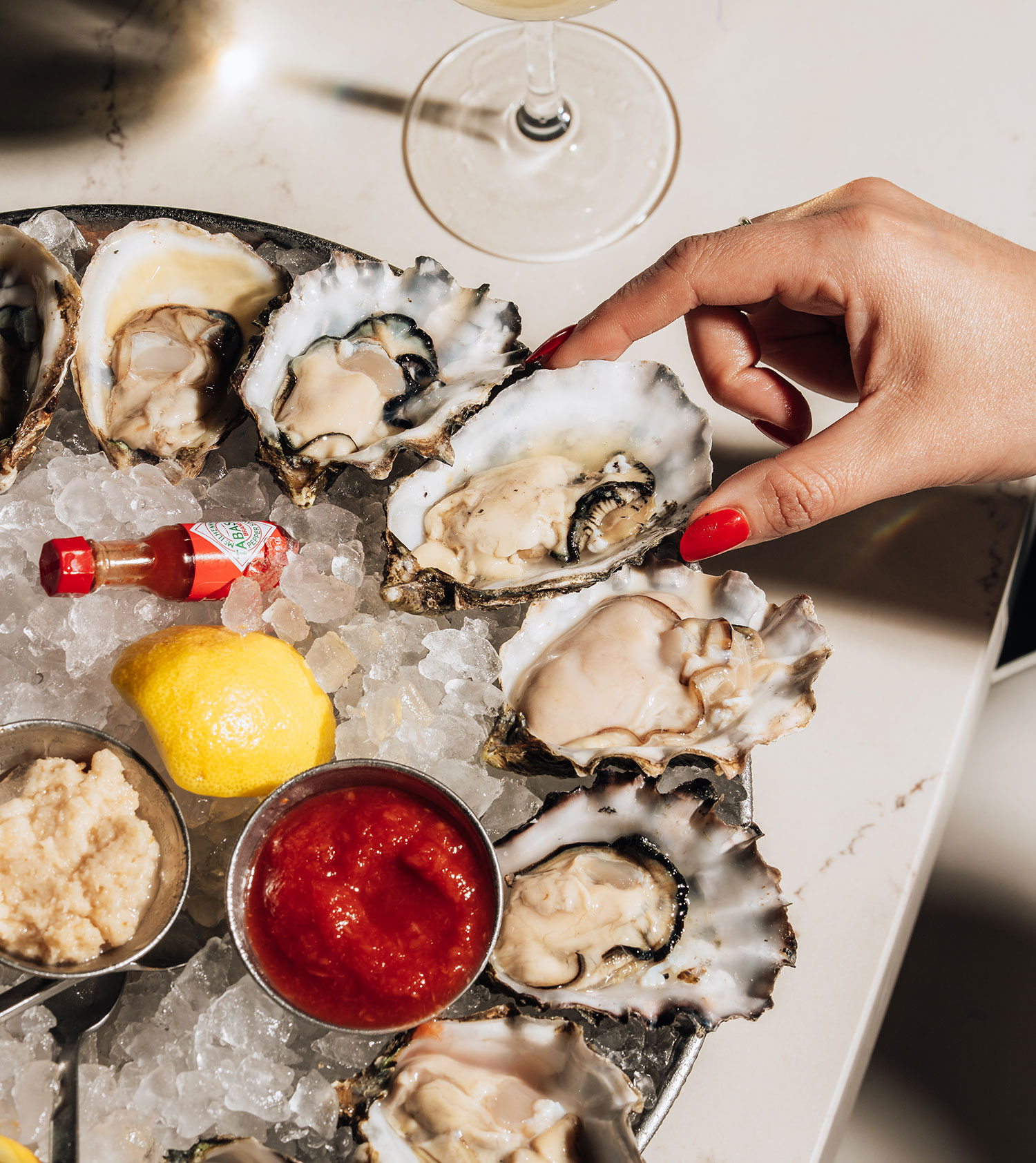 A plate of oysters with lemon and sauce in the middle. One oyster is being picked up by a hand with red nails.