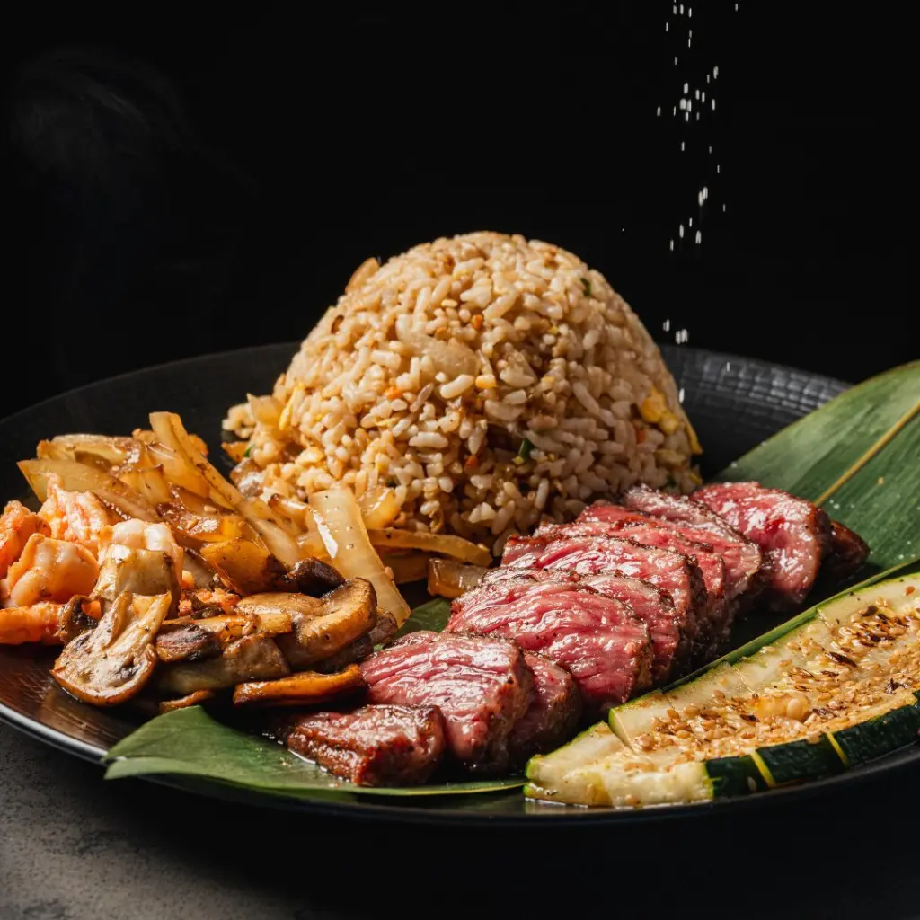 A plate of cooked steak, rice, onions and mushrooms with salt being sprinkled over