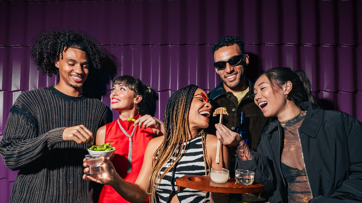 A group of happy young people stood in front of a purple wall eating with chopsticks.